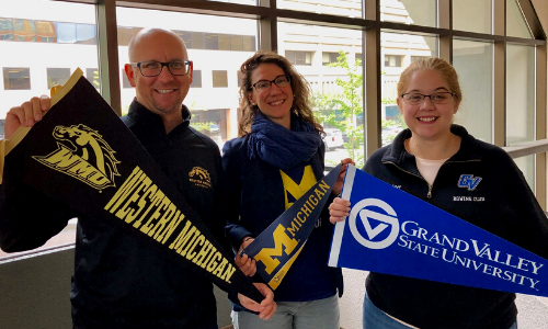 Three people (one man and two women) smiling and holding up university penents